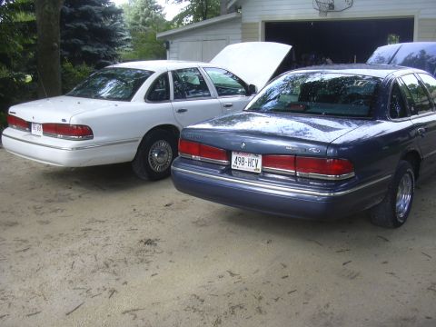 1997 Ford Crown Victoria - Two Ford Crown Victorias, a white and a blue model, are parked in front of a garage with their hoods open.