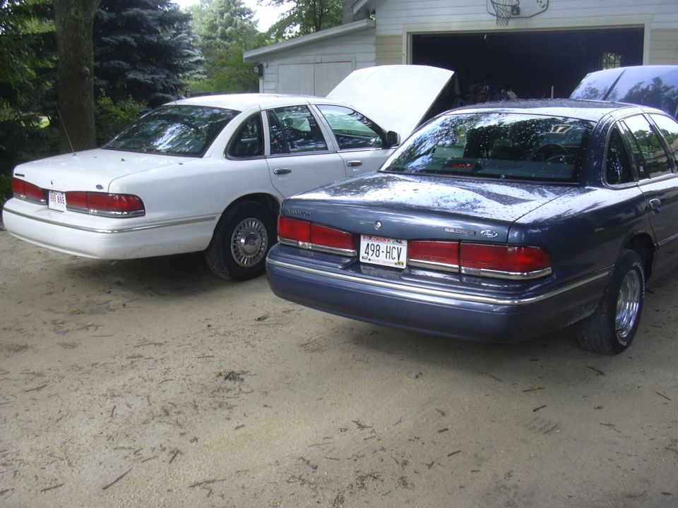 White and blue Ford Crown Victoria sedans parked with hoods up in front of a garage