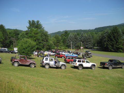 1991 Ford Bronco - A gathering of various SUVs and trucks are parked on a grassy field, with a backdrop of lush green trees and rolling hills under a clear bl…