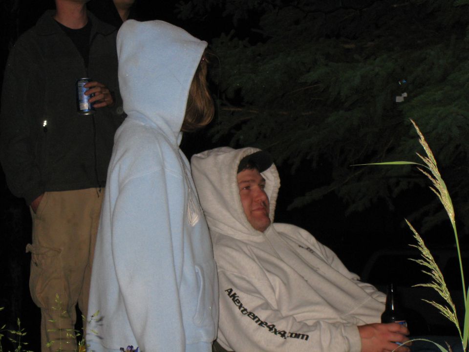 People in hoodies gathered outdoors at night, with a beer can and a bottle visible