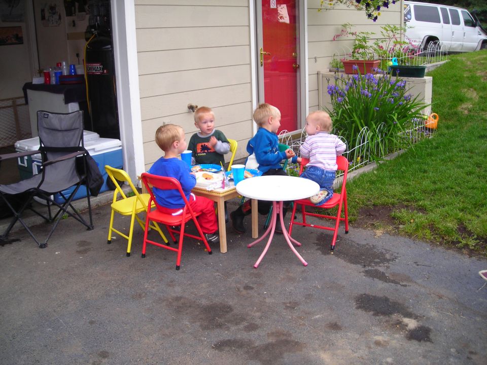Four young children sit at a small table with snacks and drinks outside a garage