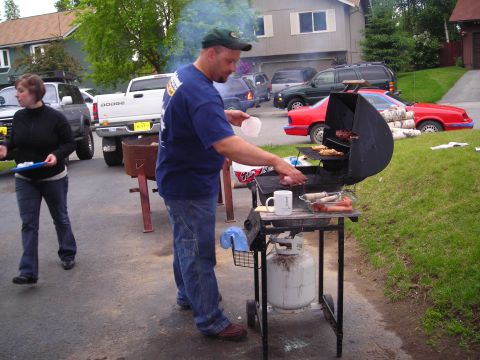 2003 Jeep TJ - A man grills food in a driveway while a woman stands nearby, with several vehicles parked in the background.