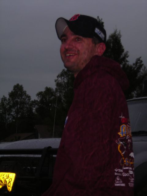 2003 Jeep TJ - A man in a maroon hoodie and baseball cap is pictured outdoors with trees and a cloudy sky. A license plate is partially visible.