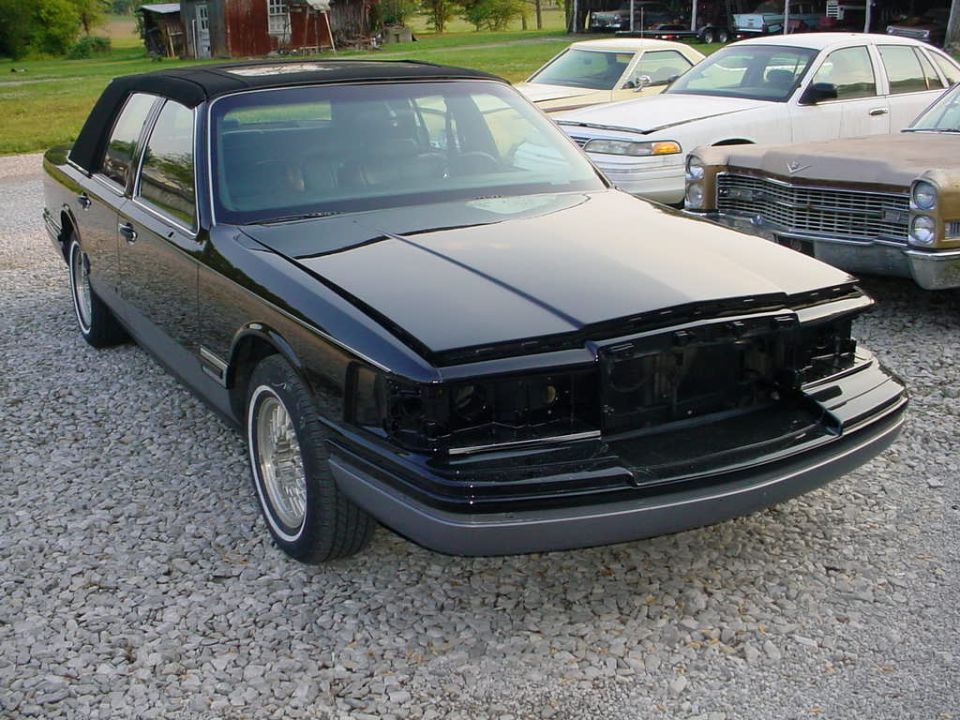 Black sedan missing grille and headlights, parked on gravel with other cars