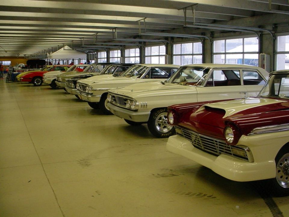 Several classic cars parked in a row inside a garage. The closest car is red and white with a scoop on the hood
