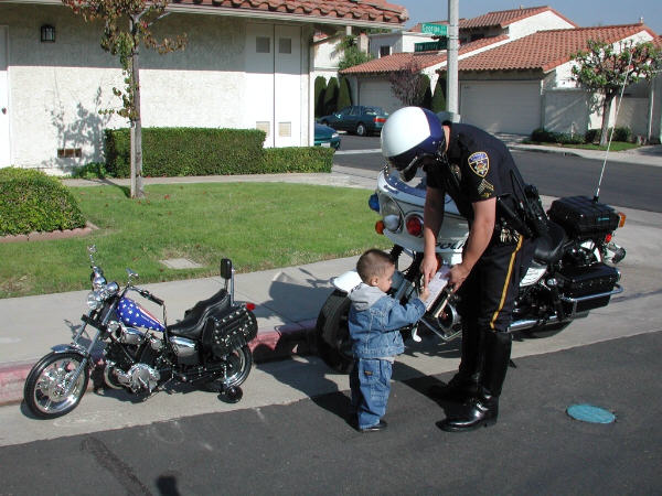 Police officer on a touring motorcycle bends to greet a toddler standing beside a small custom chopper