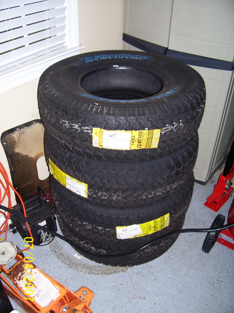 1979 Ford Bronco - Four Pro-Comp tires are stacked in a garage, with a floor jack and other equipment visible nearby.