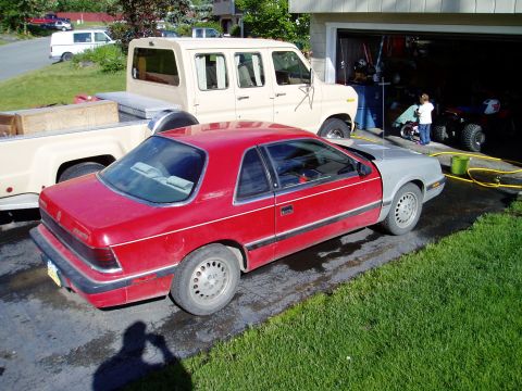 2000 Ford E350 - A red coupe with a mismatched grey hood sits in a driveway next to a beige van and pickup truck.