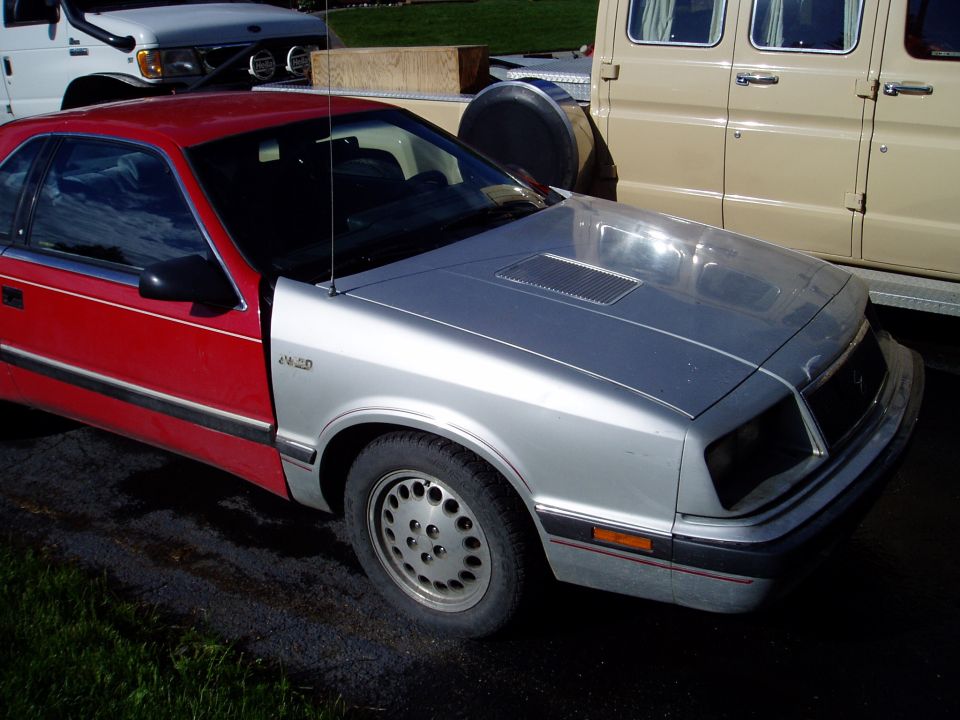Red and silver coupe with aftermarket hood scoop parked next to a beige van and white truck
