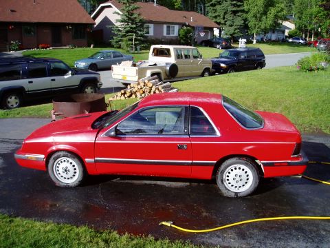 2000 Ford E350 - A red coupe is parked in a driveway with a yellow hose nearby. Other vehicles and houses are visible.