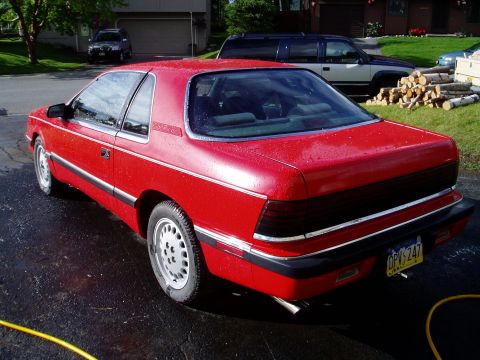 2000 Ford E350 - Red coupe with chrome trim and a black rear panel, parked on a wet driveway. Other vehicles and a pile of logs are in the background.