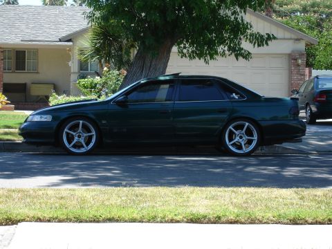 1993 Ford Taurus - A dark green 1993 Ford Taurus sedan is parked on the street in front of a house, showing its side profile and aftermarket wheels.