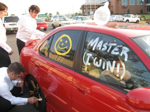 1988 Ford Bronco - People in formal attire decorate a red sedan with window paint and a large condom on the roof.