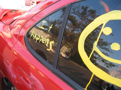 1988 Ford Bronco - A red car's rear window is decorated with yellow writing and a smiley face, obscuring the vehicle's make and model.