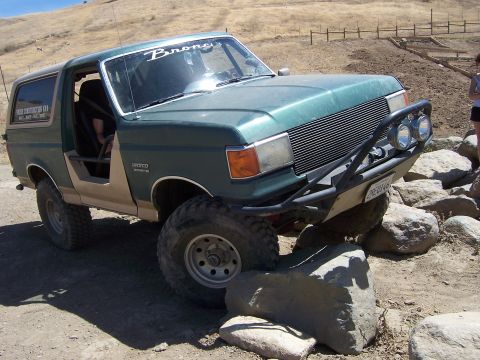1991 Ford Bronco - A green Ford Bronco is shown off-roading over large rocks on a sunny day.