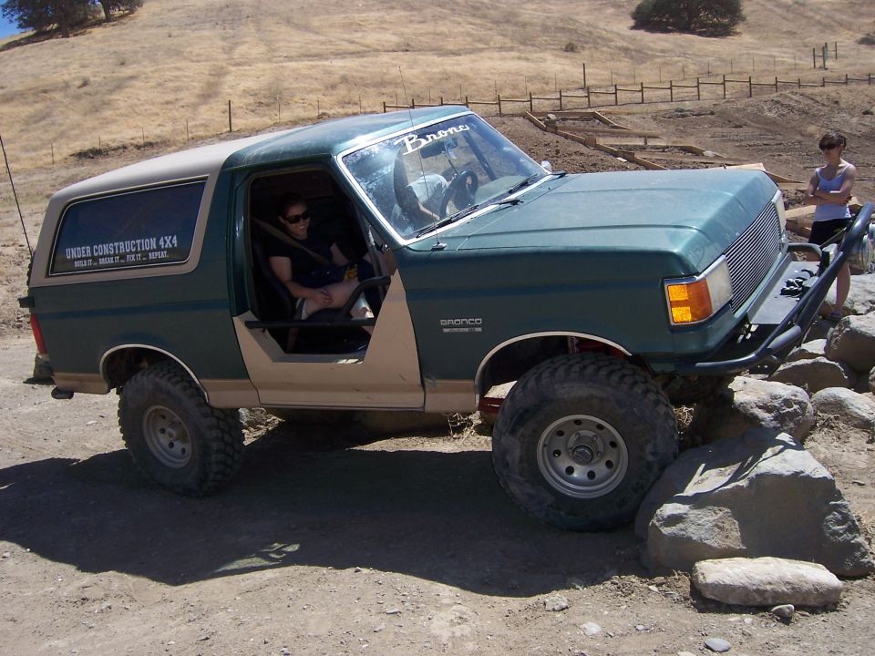 Green 1991 Ford Bronco with tan top and doors removed, driving over rocks. "UNDER CONSTRUCTION 4X4" sticker on rear quarter panel
