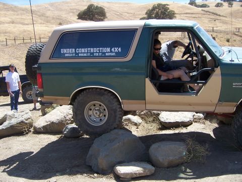1991 Ford Bronco - A green and tan Ford Bronco is shown off-roading over rocks, with a "UNDER CONSTRUCTION 4X4" sticker visible on its window.