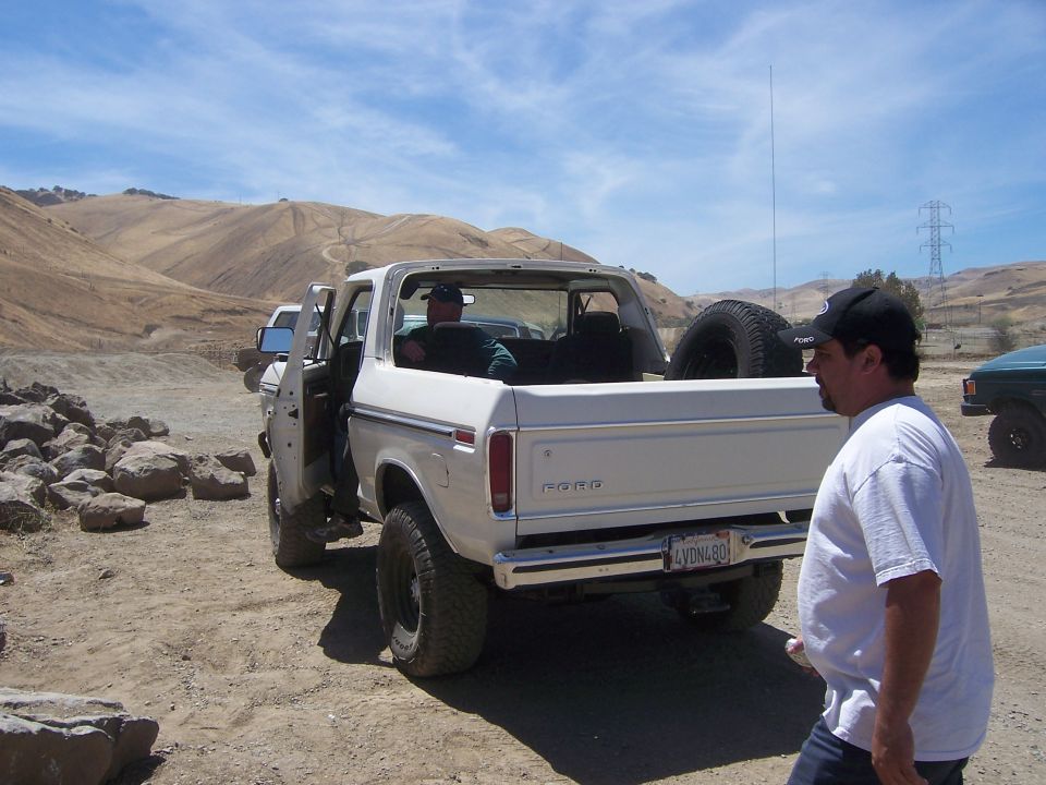 White Ford Bronco with spare tire in the bed, parked on dirt. License plate reads LVDNL80