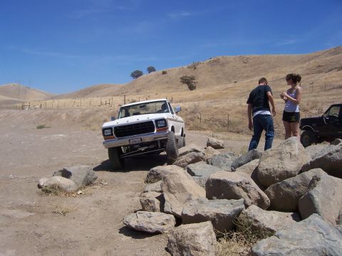 1991 Ford Bronco - A white 1979 Ford F-Series truck navigates a rocky path, with two individuals observing the off-road excursion.