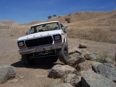 1991 Ford Bronco - White Ford truck with lifted suspension drives over rocks on a dry, hilly landscape.