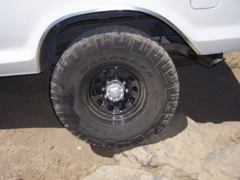 1991 Ford Bronco - A close-up view of an off-road tire and wheel on a white vehicle, positioned on rocky ground.