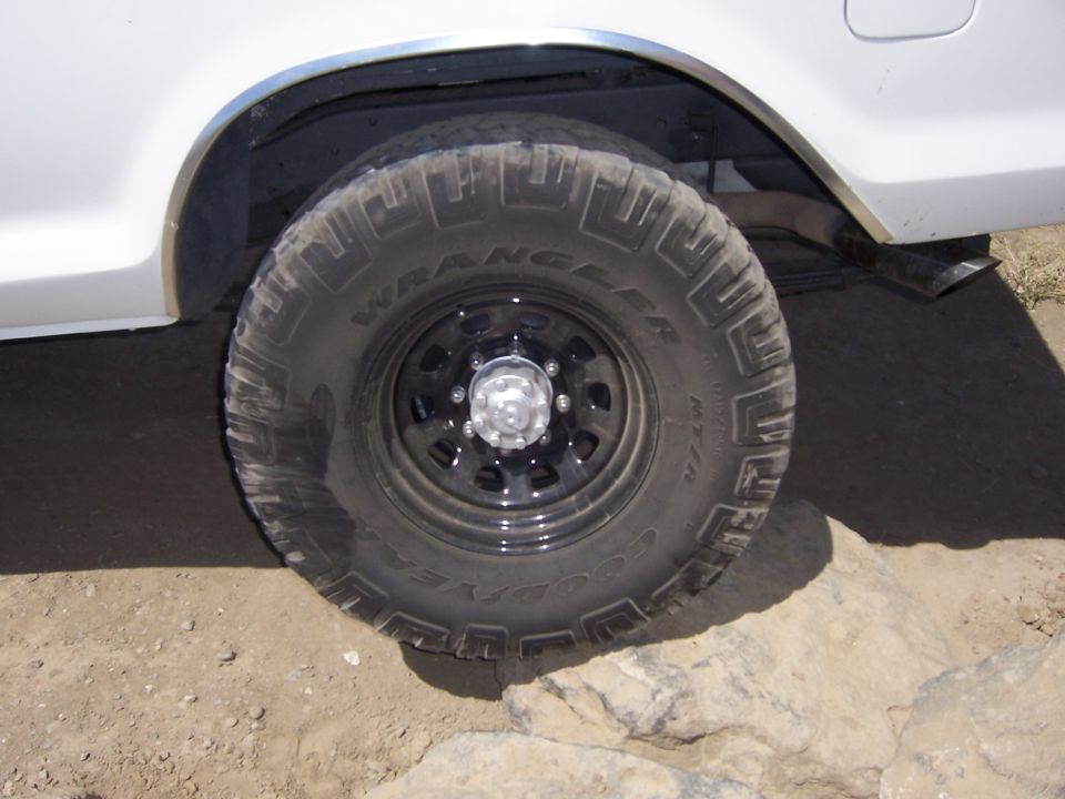 Close-up of a Goodyear Wrangler M/T tire on a black rim, mounted on a white vehicle. The tire is on rocky terrain