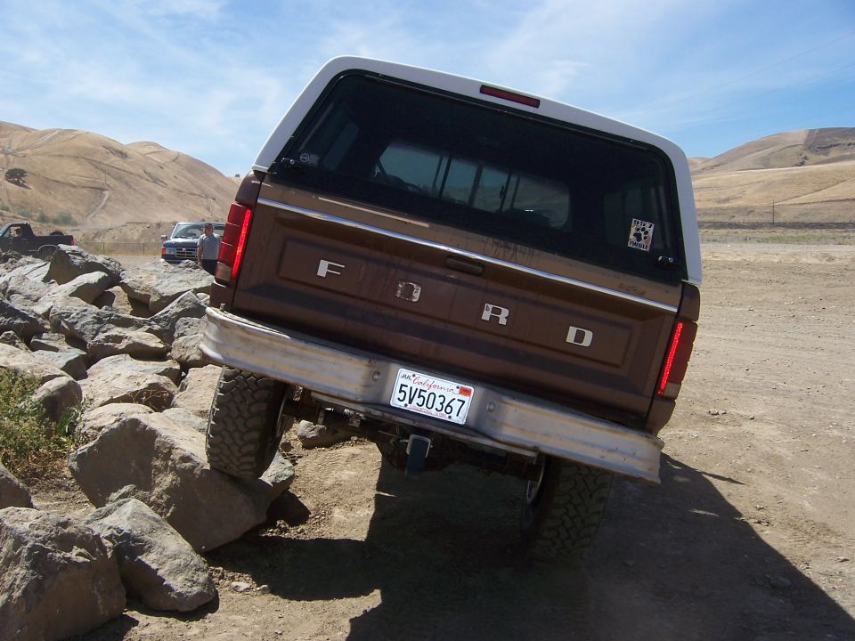 Brown 1987 Ford Bronco with white camper shell driving over rocks. California license plate 5V50367