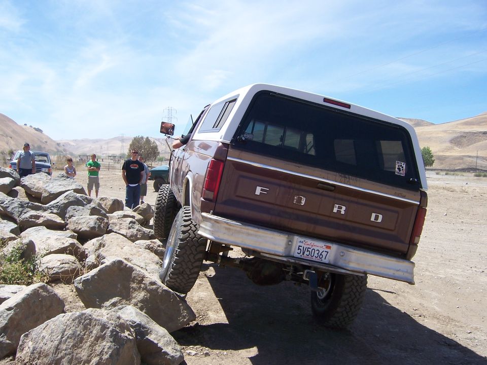 Brown Ford Bronco with white camper shell driving over rocks. Other vehicles and people in background