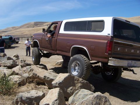 1991 Ford Bronco - A lifted Ford F-Series pickup truck navigates a rocky terrain with a white camper shell on its bed.