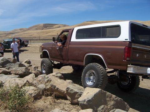 1991 Ford Bronco - A lifted brown pickup truck navigates a rocky obstacle course under a clear blue sky.
