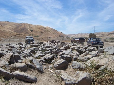 1991 Ford Bronco - Several Ford vehicles traverse a boulder-strewn path under a partly cloudy sky.