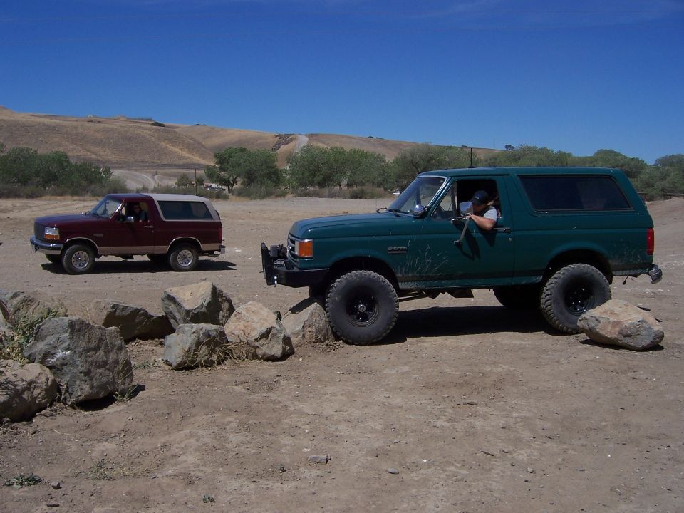 Green 1991 Ford Bronco driving over rocks, maroon Bronco behind it