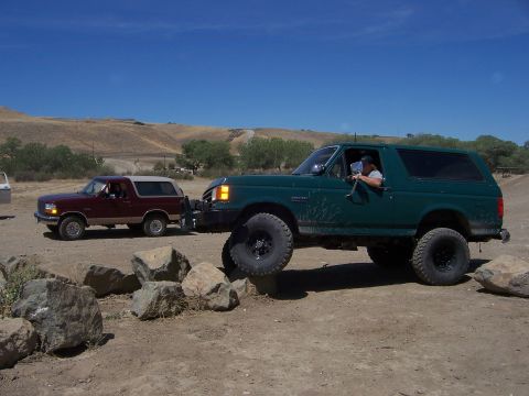 1991 Ford Bronco - Two Ford Broncos navigate a rocky off-road course under a clear blue sky.