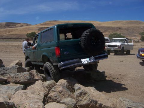 1991 Ford Bronco - A green Ford Bronco navigates a rocky terrain, with another white Ford Bronco visible in the background.