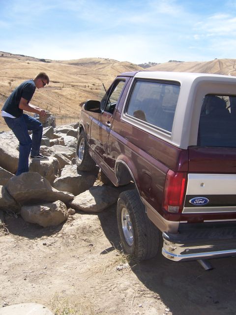 1991 Ford Bronco - A man stands on rocks next to a maroon and tan Ford Bronco on a dirt path with rolling hills in the background.