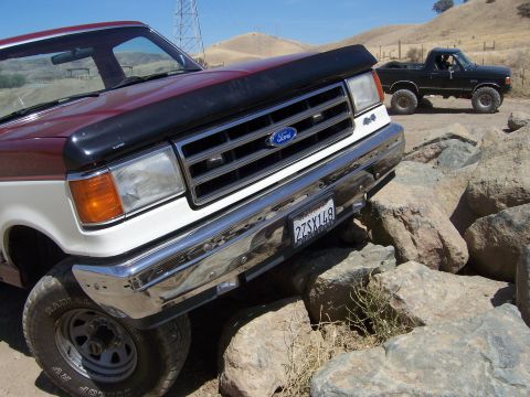 1991 Ford Bronco - A maroon and white Ford F-Series truck navigates a rocky terrain, with another black truck visible behind it.