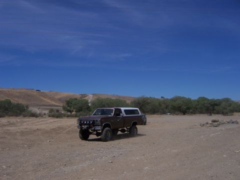 1991 Ford Bronco - Lifted pickup truck with camper shell on a dirt road under a clear blue sky.