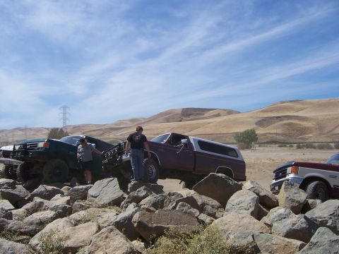 1991 Ford Bronco - Three trucks, including a maroon 1991 Ford Bronco, traverse a challenging rocky landscape under a blue sky.