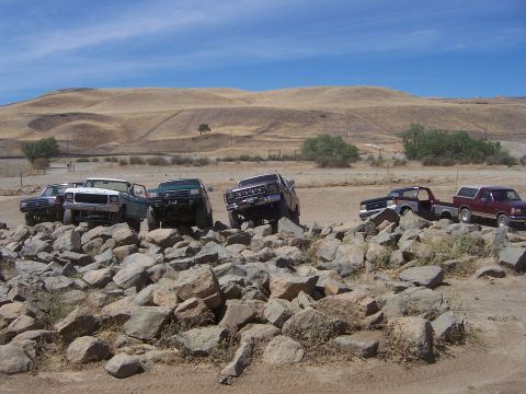 1991 Ford Bronco - Multiple trucks are shown off-roading over a rocky obstacle course on a sunny day.