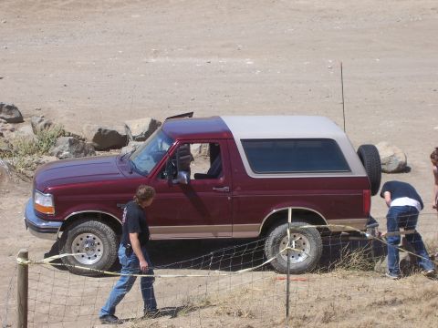 1991 Ford Bronco - A maroon Ford Bronco with a tan removable top is parked on a dirt lot with two people working near its rear bumper.