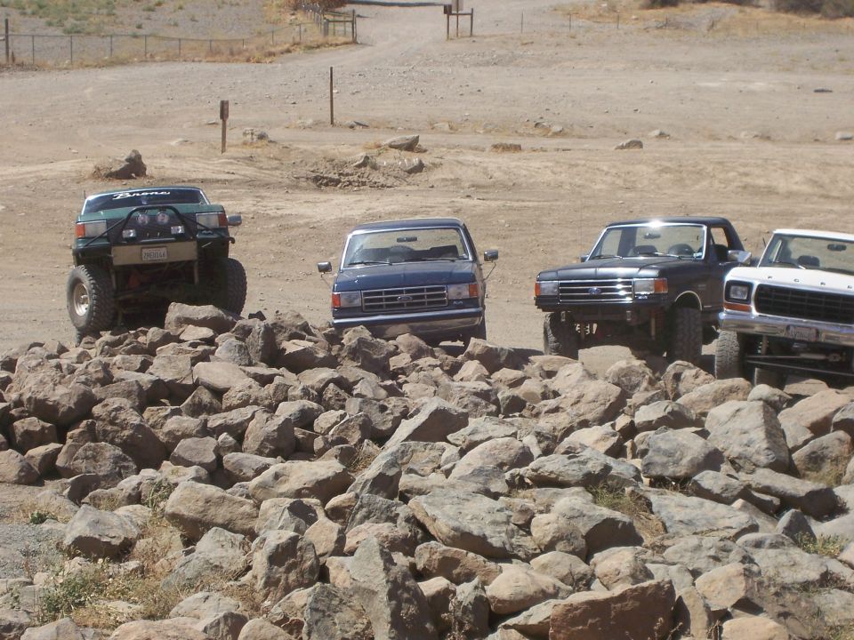 Four Ford trucks and SUVs on a rocky terrain, front view. One green Bronco is lifted with off-road tires