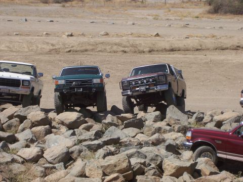 1991 Ford Bronco - Several trucks and SUVs are shown off-roading on a rocky trail, with a maroon Ford Bronco in the foreground.