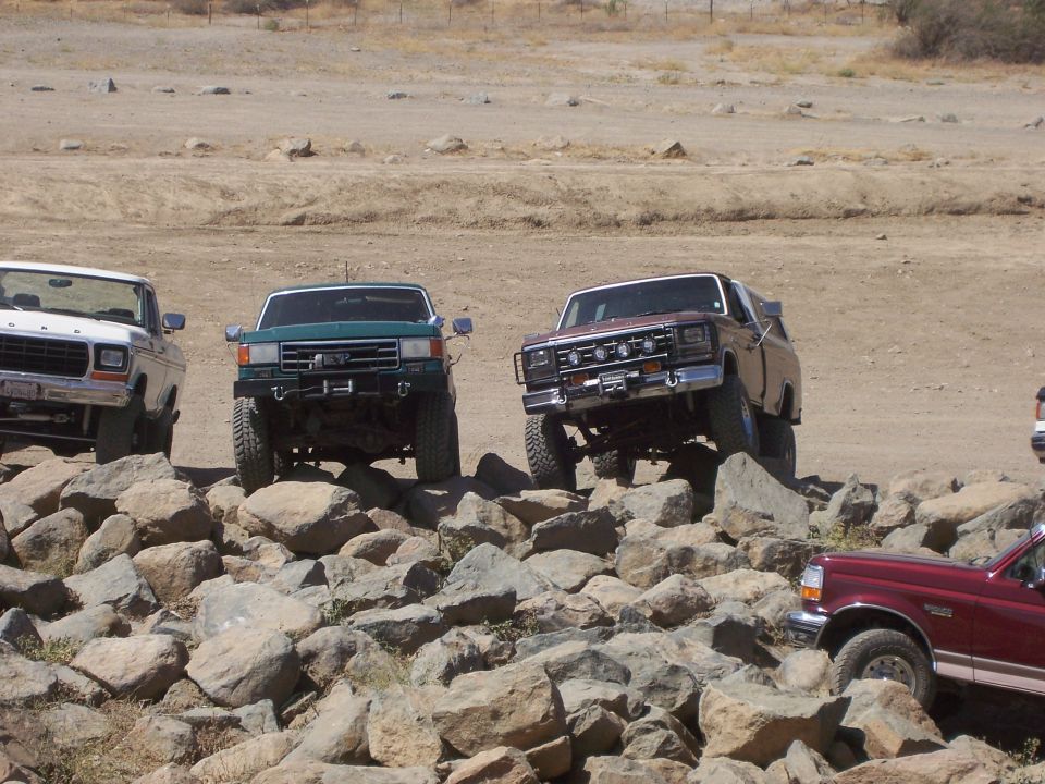 Four-wheel drive trucks and SUVs navigating a rocky obstacle course. The lead vehicle is a maroon Ford Bronco