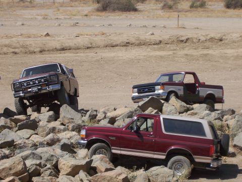 1991 Ford Bronco - Multiple Ford Broncos and an F-150 are shown off-roading over rocks on a sunny day.