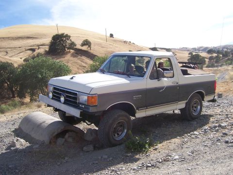 1991 Ford Bronco - A two-tone Ford Bronco navigates an off-road obstacle on a sunny day.