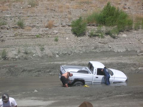 1985 Ford Bronco - Two people attempt to free a white pickup truck that is stuck deep in mud.
