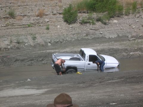 1985 Ford Bronco - Two people attempt to free a white pickup truck that is stuck in mud and water.