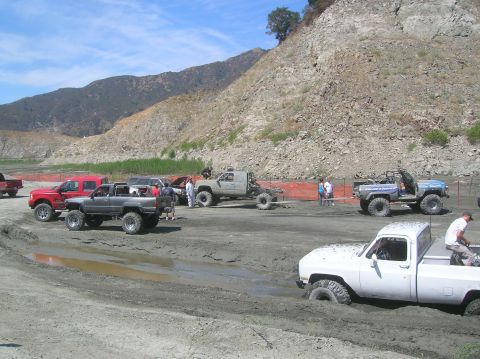 1985 Ford Bronco - Several off-road vehicles are stuck in the mud near a rocky embankment on a sunny day.