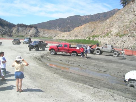1985 Ford Bronco - Several off-road vehicles navigate a muddy terrain, with one red pickup truck deeply mired in a water-filled rut.