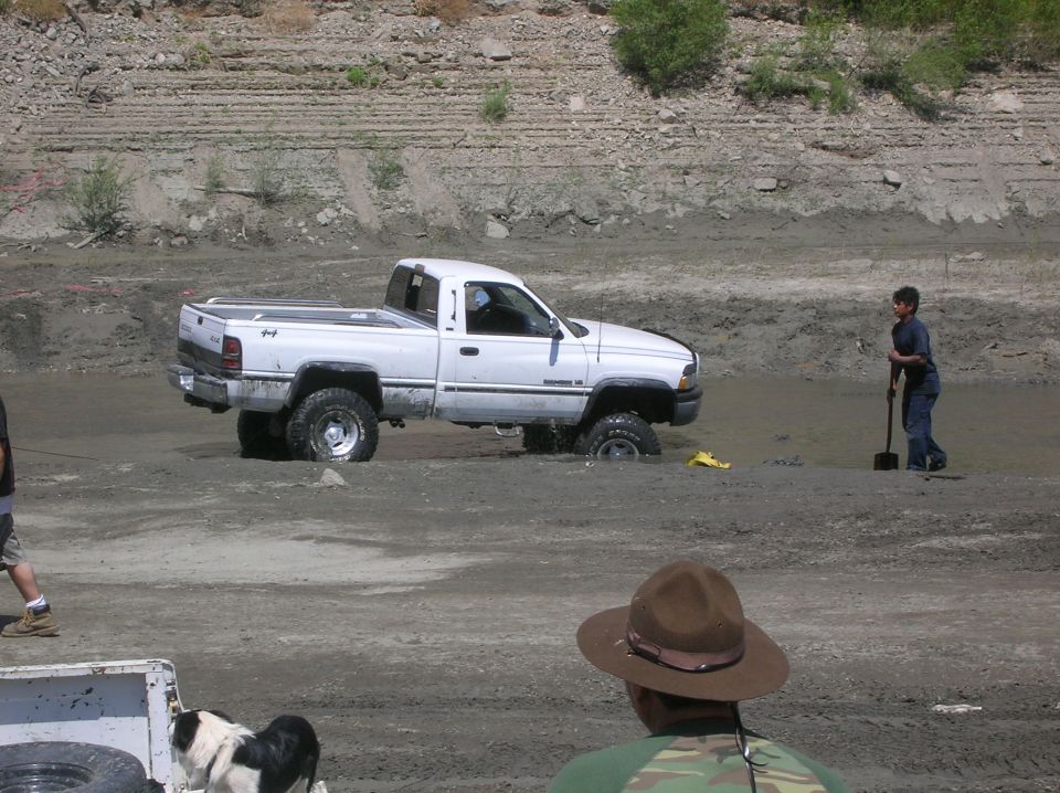 White pickup truck stuck in mud with a person holding a shovel nearby